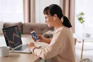 Smiling business woman trader analyst looking at laptop monitor, holding smartphone, wearing earphones. Investor broker analyzing indexes, trading online investment data on stock market graph at home<br>investiranje, mladi, studenti, ulaganje, učenje, burze