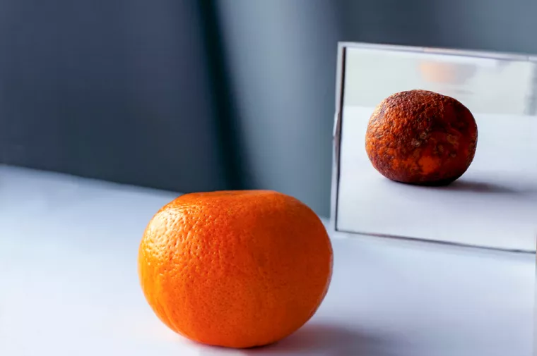 А fresh orange on the table is reflected in the mirror as rotten фтв spoiled fruit. Conceptual photo about young and old age, inner beauty, loneliness, lies.<br>trulo voće, truljenje, naranča, ogledalo
