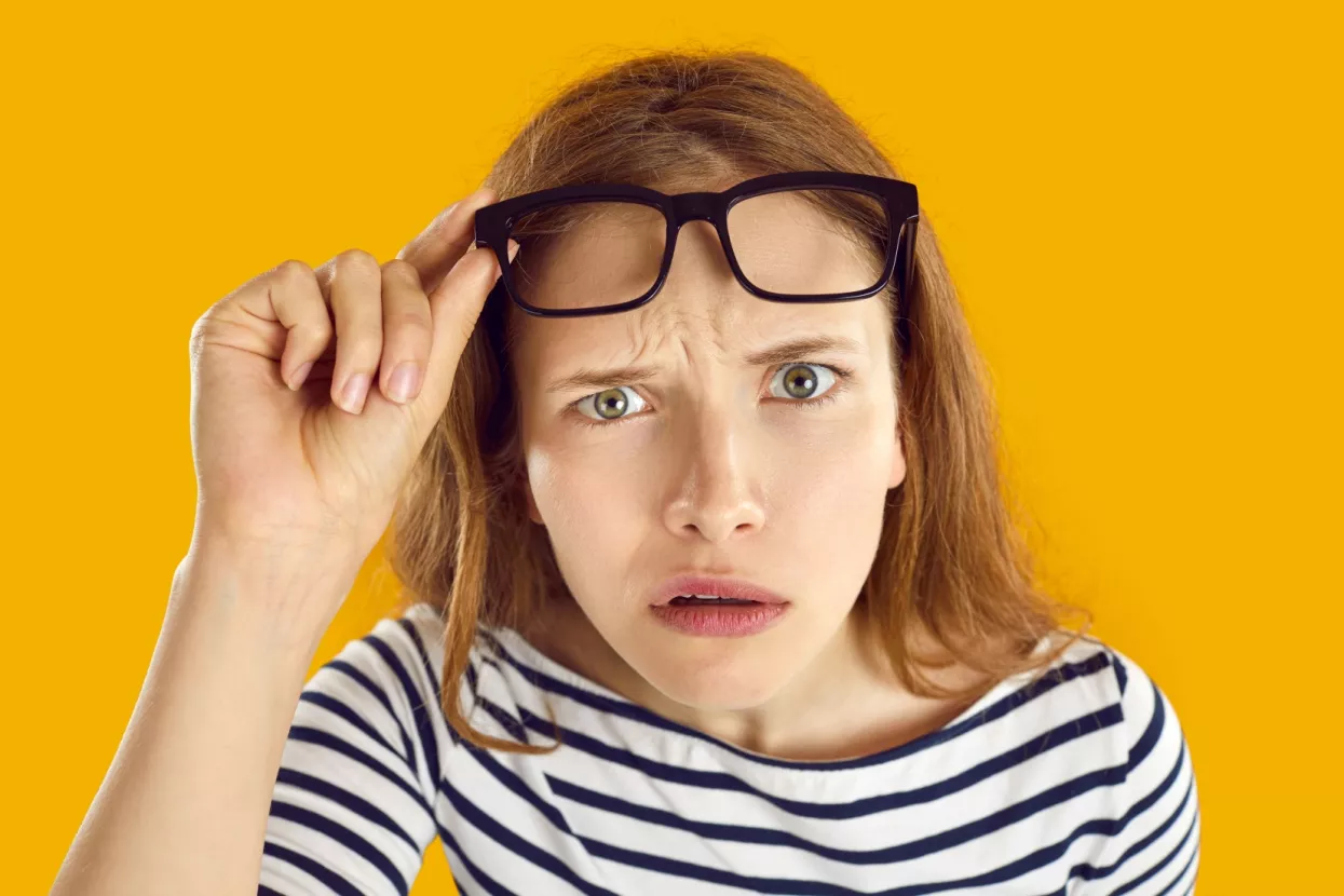 Confused woman looks at you with suspicious, doubtful and incredulous expression on her face. Close up of skeptical and suspicious young woman looking out from under glasses on orange background.
