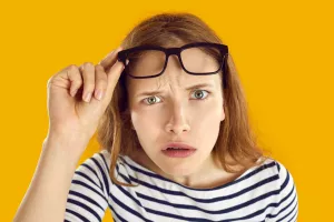 Confused woman looks at you with suspicious, doubtful and incredulous expression on her face. Close up of skeptical and suspicious young woman looking out from under glasses on orange background.