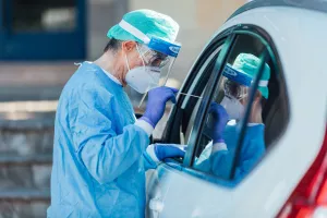 Medical personnel wearing a PPE, performing PCR with a swab in their hand, on a patient inside his car to detect if he is infected with COVID-19