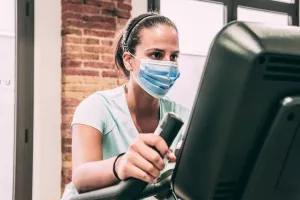 Young brunette woman does static cycling at the gym. She's wearing a mask.