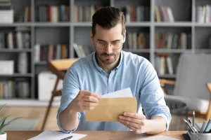 Serious businessman sit at workplace office desk holding envelope take out letter feels interested read business news, got invitation, learns bank statement information. Postal correspondence concept