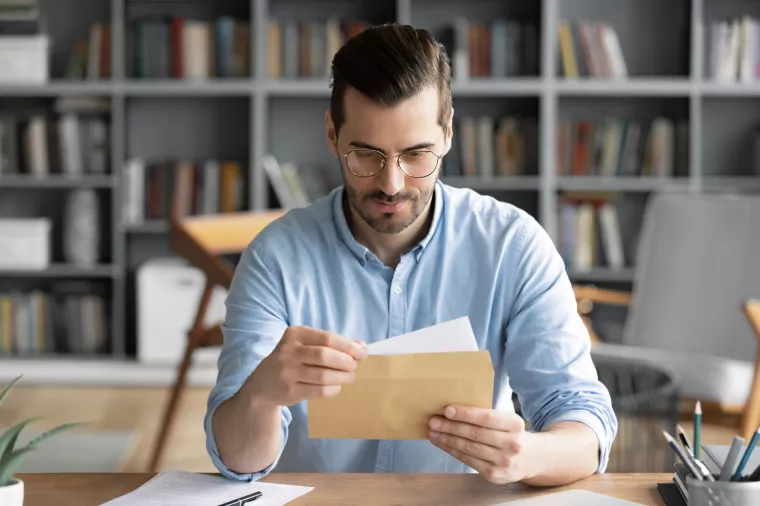 Serious businessman sit at workplace office desk holding envelope take out letter feels interested read business news, got invitation, learns bank statement information. Postal correspondence concept