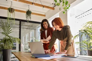 Two smiling professional female partners or coworkers, happy business women entrepreneurs working together in office looking at laptop using computer writing notes standing at work desk.