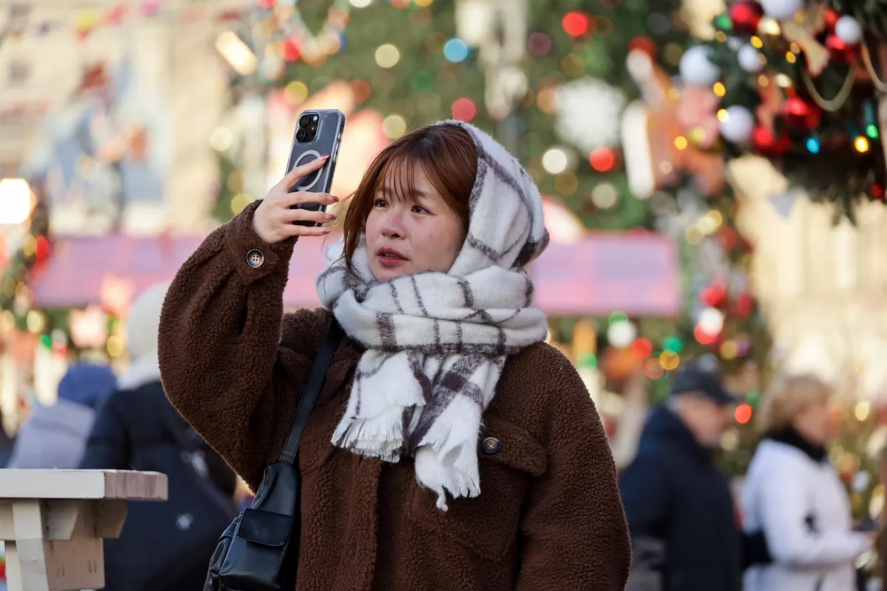 Moscow, Russia - December 5, 2024Asian girl taking pictures on smartphone camera standing on New Year trees background, tourist in winter cityAzija, azijatkinja, turisti, turistkinja, turizam